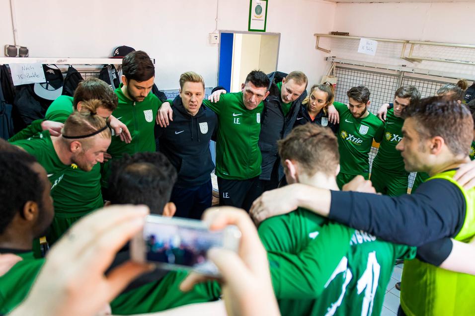 Mannschaftsbesprechung in der Umkleide mit Spielern in grünen Trikots und Trainer, alle im Kreis versammelt.