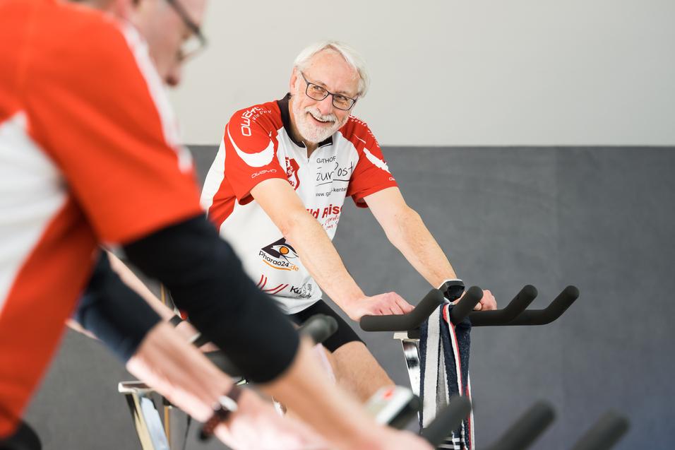 Älterer Mann mit Brille und grauen Haaren lächelt auf einem Spinning-Bike in einer Gruppe von Radfahrern.