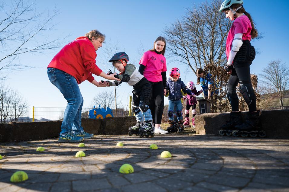 Kinder üben auf Rollschuhen mit Hilfe eines Erwachsenen auf einem gepflasterten Platz, umgeben von Tennisbällen.