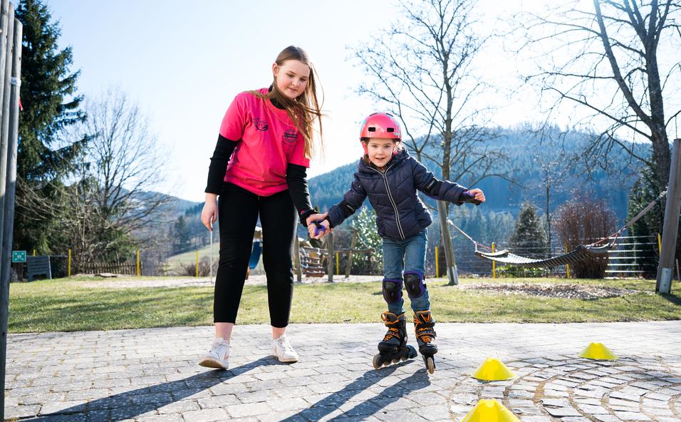 Ein Mädchen übt mit einer Trainerin das Inline-Skaten auf einem gepflasterten Platz, umgeben von Bäumen und Hügeln.