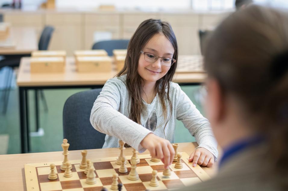 Mädchen mit Brille zieht eine Schachfigur auf einem Schachbrett in einem Klassenraum. Holzschachfiguren im Vordergrund.