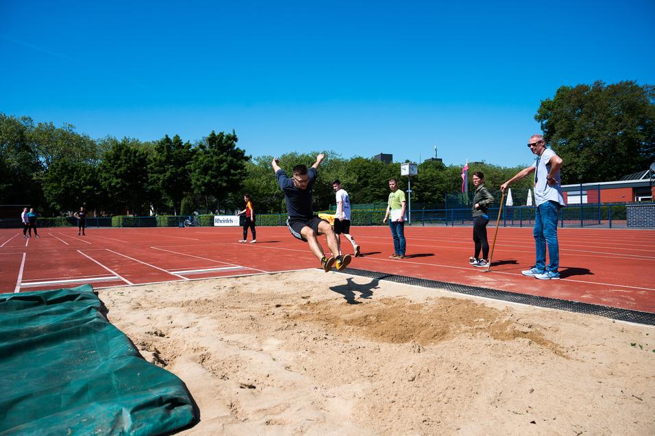 Sprinter springt vom Absprungbrett in die Sandschicht einer Weitsprunganlage, während Zuschauer am Rand stehen.