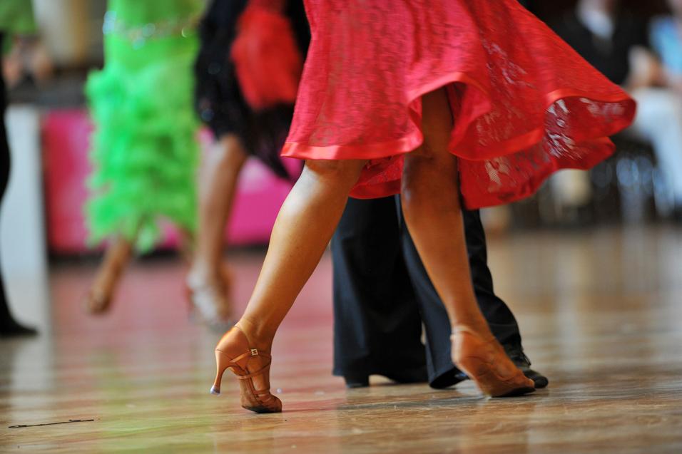 Tanzende Beine in eleganten Schuhen, während sich ein rotes Kleid dreht, vor unscharfem Hintergrund.