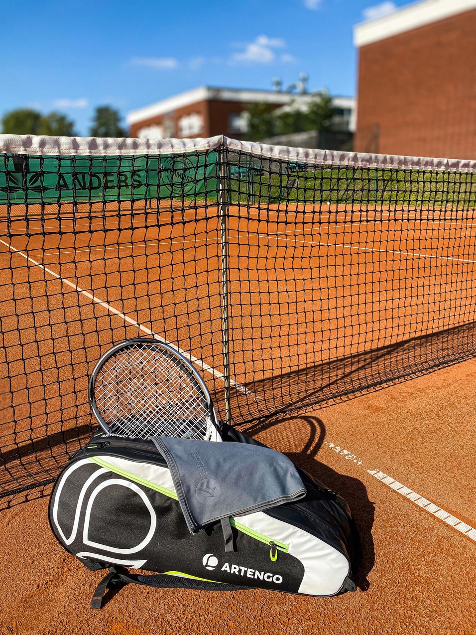 Tennisplatz mit Netz, einer ARTENGO-Tasche und einem Schläger im Vordergrund auf dem rotem Sand.