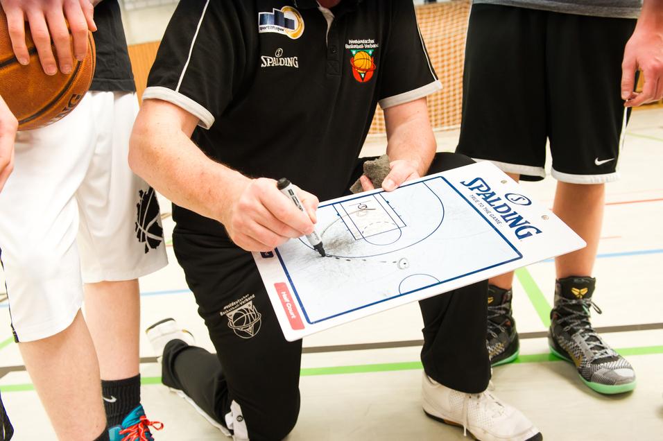 Trainer kniet mit Taktiktafel und zeichnet Spielzüge, während Spieler im Hintergrund stehen und einen Basketball halten.