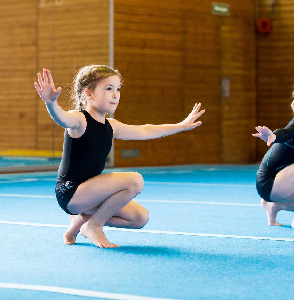 Zwei Mädchen in Gymnastikanzügen üben auf einer blauen Matte, eine kniet mit ausgestreckten Armen.