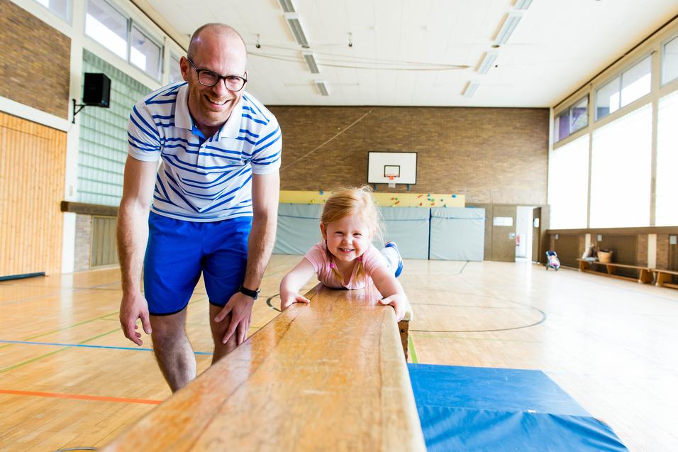 Mann lehnt am Schwebebalken, während ein lächelndes Mädchen darauf liegt, in einer hellen Sporthalle.