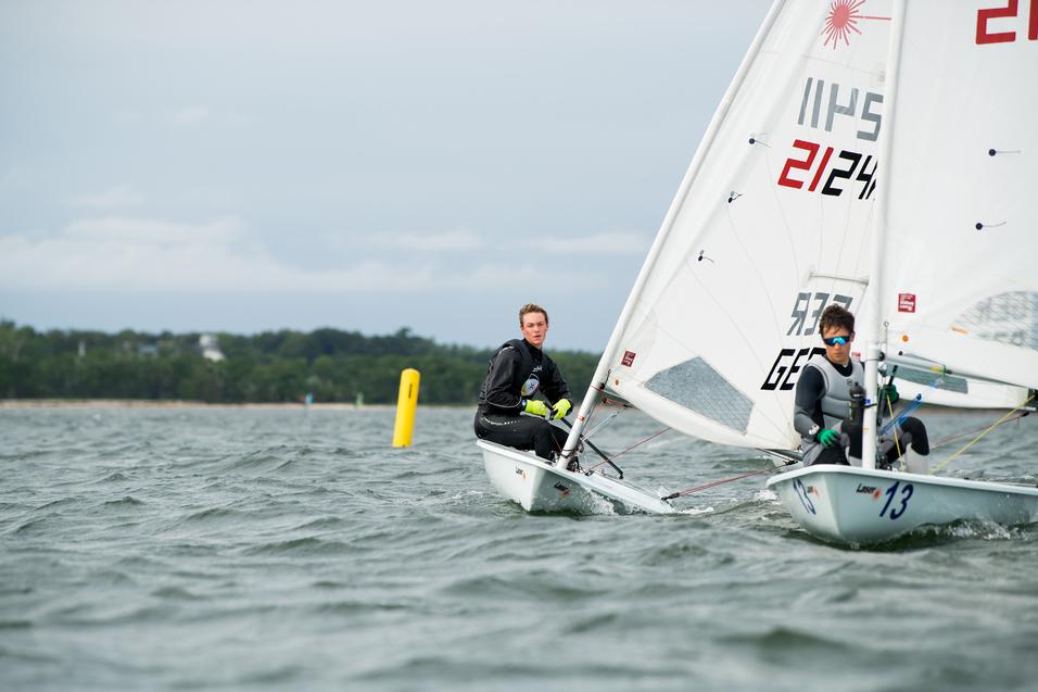 Zwei Segler in einem Boot gleiten über das Wasser, gelbe Boje im Hintergrund, bewölkter Himmel.