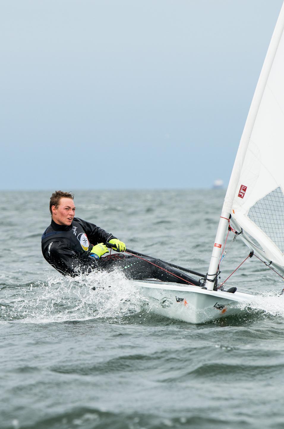 Segler in Schwarz, mit gelben Handschuhen, steuert ein kleinbootiges Segelboot über die Wellen eines Gewässers.